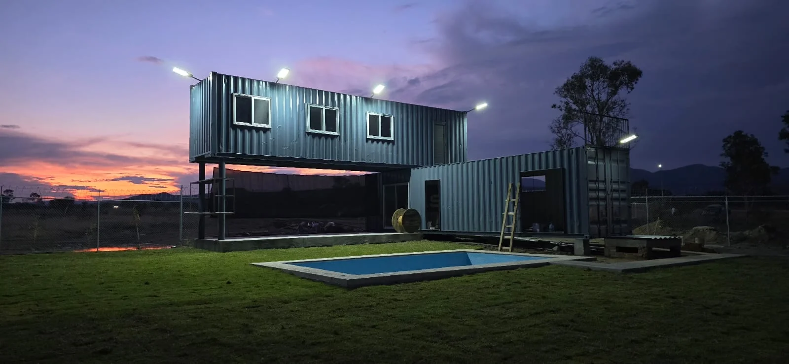 Casa de dos niveles construida con contenedores, iluminada al atardecer junto a una alberca
